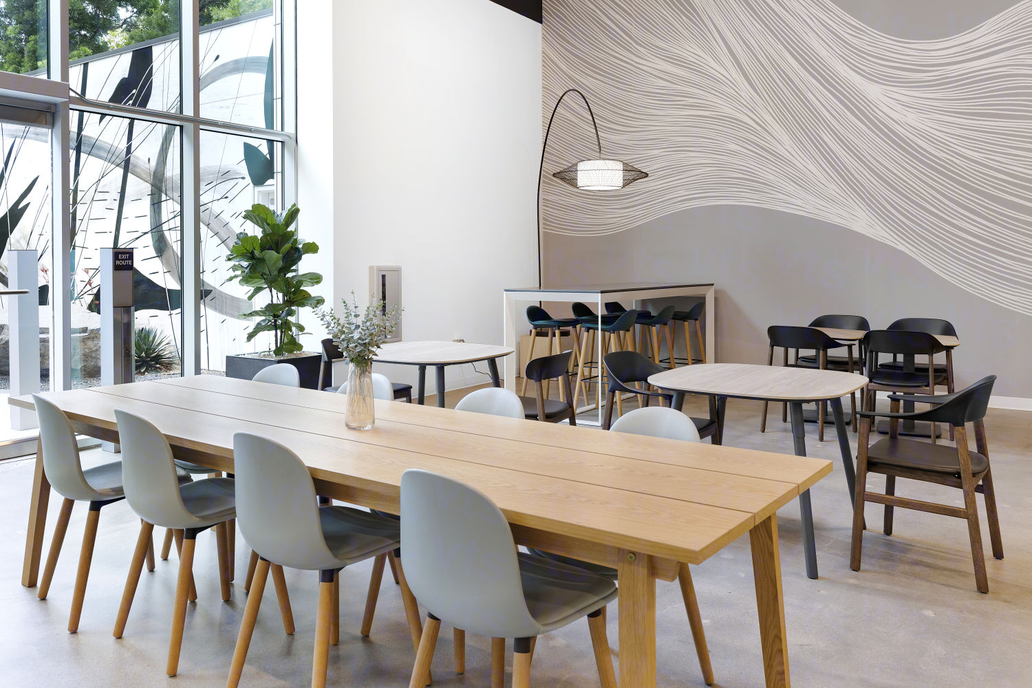 Multiple square and long tables with surrounding chairs in an open, inviting space next to wall height windows