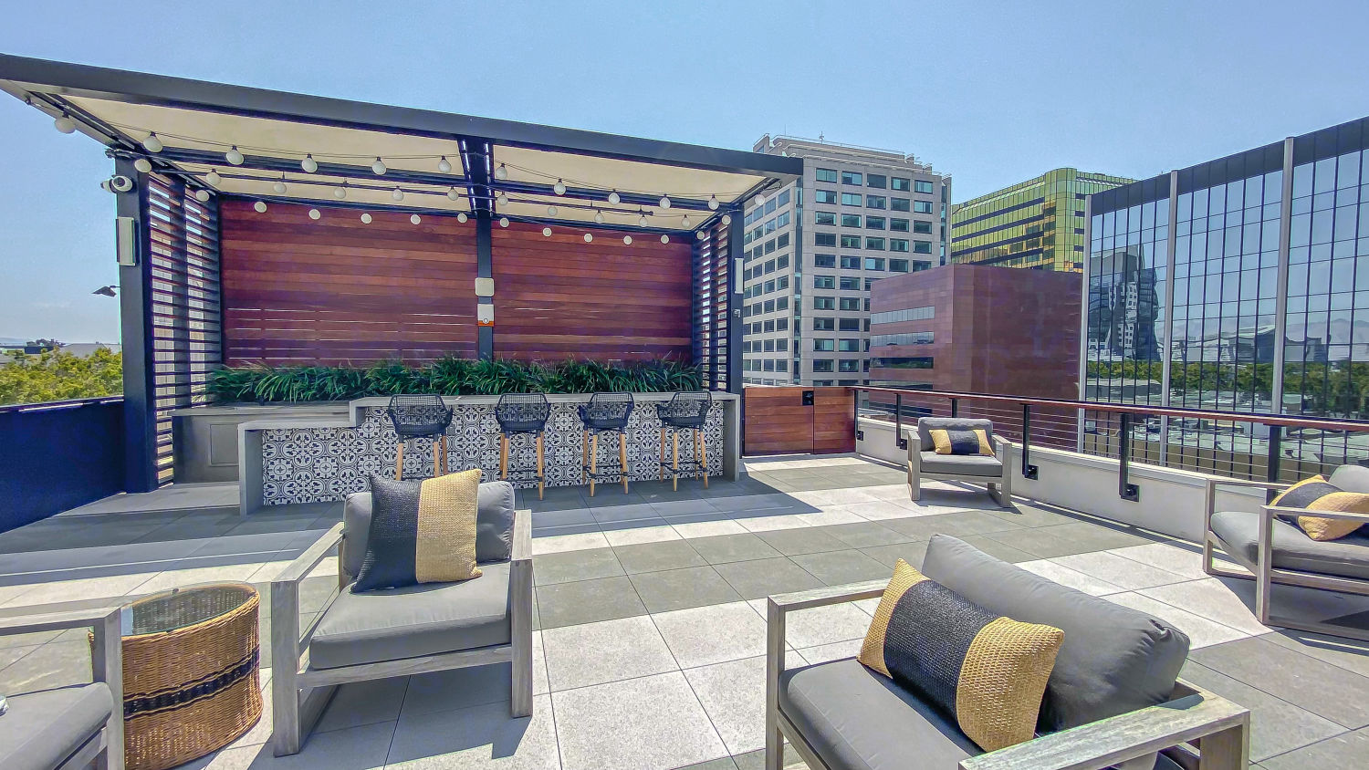 A rooftop patio consisting of a partially covered bar table and barstools surrounded by cushion lounge chairs on a clear sunny day