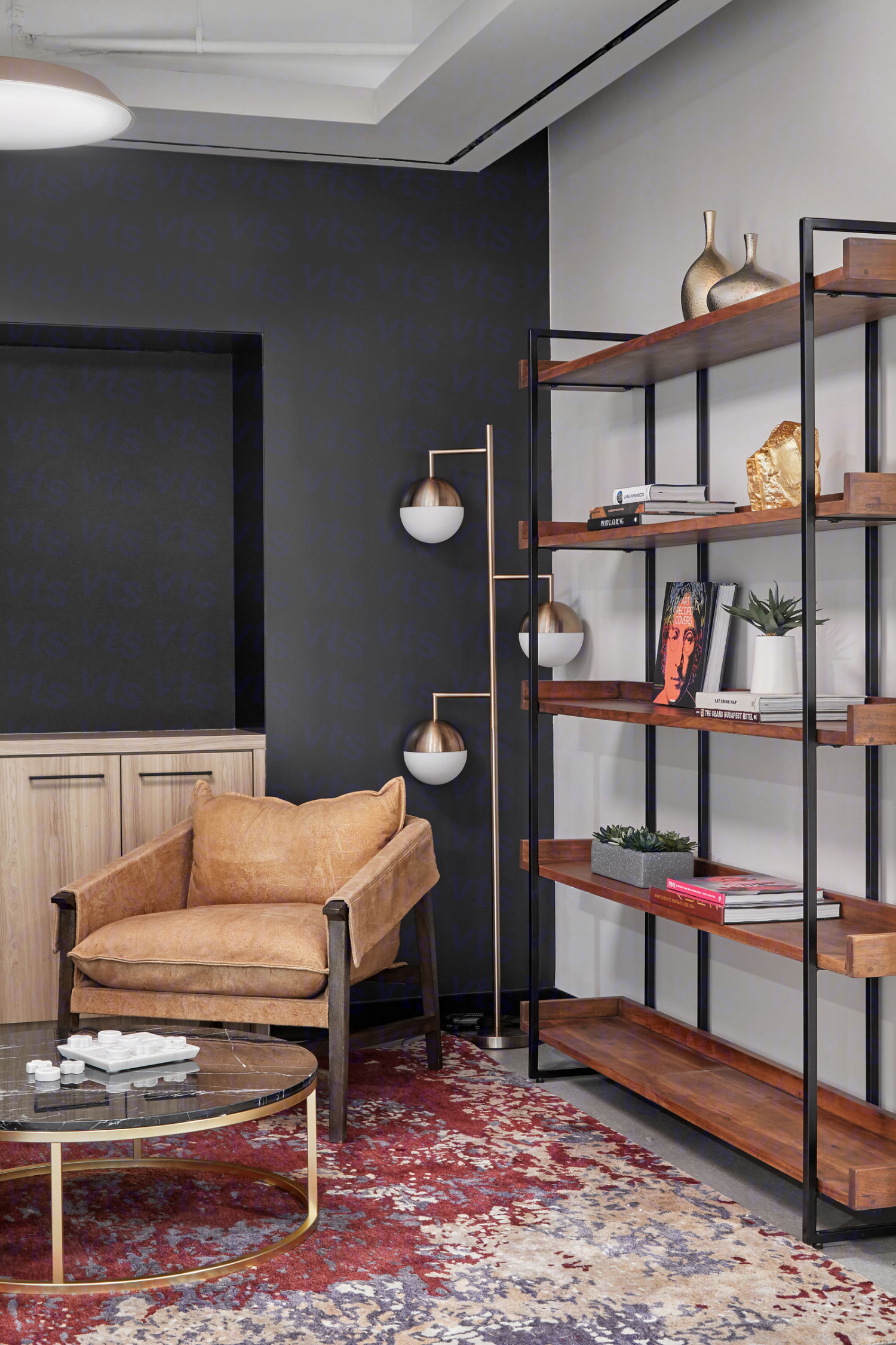 A corner of a room with a tan reading chair and a low marble coffee table against a dark painted wall adjacent to a dark wooden bookshelf with assorted books and decorations