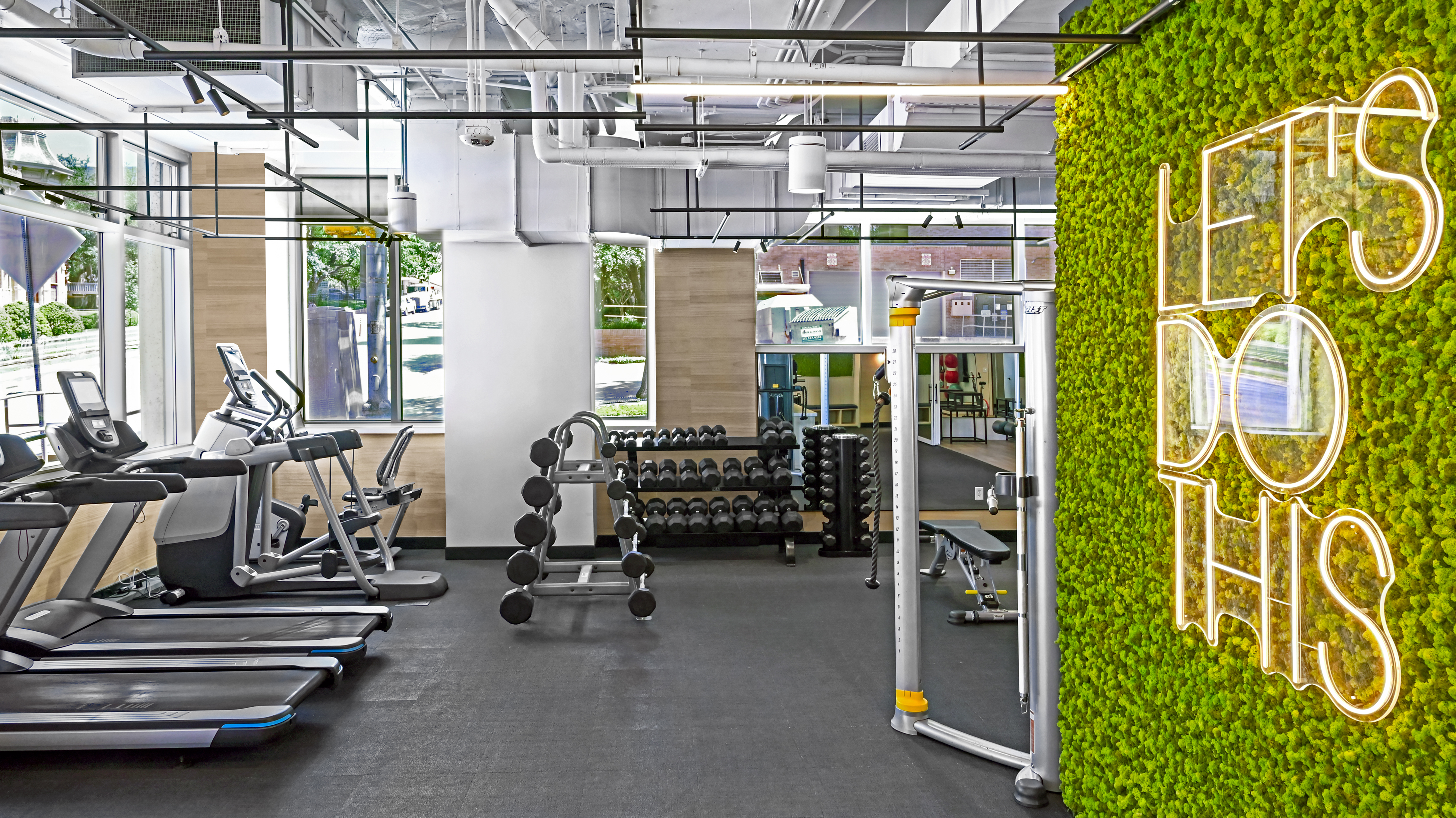 Modern fitness center featuring a row of treadmills, free weight racks, and a vibrant green moss wall with a neon sign that reads 'LET'S DO THIS.' loading=